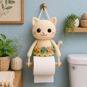 May include: A cream crochet cat toilet roll holder with a floral skirt. The cat has black eyes, pink cheeks, and whiskers. The toilet roll is held by a wooden dowel. The holder is suspended from a brass hook.