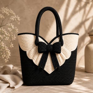 Puede incluir: Un bolso tipo tote de ganchillo en color negro y crema con un panel superior festoneado en color crema y un lazo de ganchillo negro prominente centrado en la parte delantera. El bolso tiene dos asas de ganchillo negras y una base rectangular.