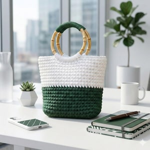 May include: A handmade crochet tote bag with a white upper section and a dark green lower section. The bag features bamboo handles with a green accent and a white handle wrap. The bag is sitting on a white surface.