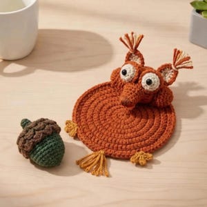 May include: A handmade crochet squirrel coaster in shades of orange, with large eyes and a long snout. A separate green and brown crochet acorn sits nearby. The coaster has small yellow tassels and is on a light wood surface.