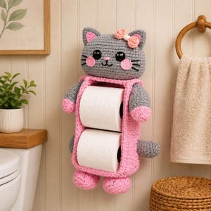 May include: A gray and pink crochet cat-shaped toilet paper holder. The holder has a cat face with a pink bow, holding two rolls of white toilet paper. The cat has pink paws and ears. The holder is mounted on a white wall.