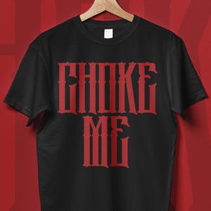 Peut inclure: T-shirt noir avec l'inscription "CHOKE ME" en rouge, dans une police gothique. Le t-shirt est suspendu à un cintre en bois sur un fond rouge. Le texte est centré sur le devant du vêtement.