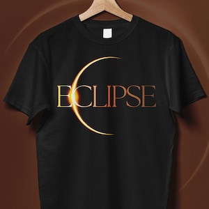 Op de afbeelding: Zwart T-shirt met het woord "ECLIPSE" in een gouden kleurovergang en een gouden halve maan. Het T-shirt hangt op een houten hanger tegen een bruine achtergrond.