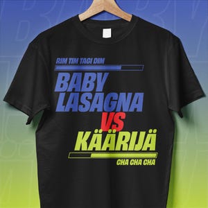 Könnte beinhalten: Schwarzes T-Shirt mit dem Text "RIM TIM TAGI DIM" über "BABY LASAGNA VS KÄÄRIJÄ" und "CHA CHA CHA" darunter. Der Text ist in Blau, Rot und Gelb gehalten. Das Shirt wird auf einem Holzbügel präsentiert.