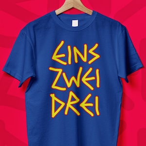 May include: A blue t-shirt with the German words "EINS ZWEI DREI" in yellow, stylized lettering. The shirt is hanging on a wooden hanger against a red background. The text translates to "one two three".
