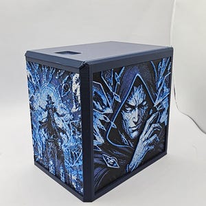 May include: A dark blue rectangular box featuring detailed 3D-printed panels on the sides. One side displays a hooded figure with sharp facial features, while the other side shows a silhouette of a figure standing amidst swirling blue energy patterns.