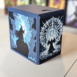 May include: A dark blue box with fantasy art on the sides. One side features a wizard with a staff and lightning, the other a circular design with a robed figure. The box has a slot on top.