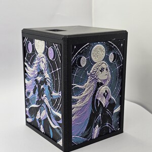 May include: A black rectangular box featuring two side panels with detailed illustrations of a long-haired figure in dark armor against a backdrop of celestial moon phases in shades of purple, blue, and white, with a square opening on the top surface.