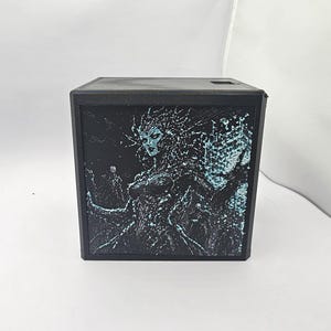 May include: A black square box featuring a detailed lithophane panel on the front. The panel displays a high-contrast, textured image of a figure with spiky hair and intricate patterns in shades of black and light blue, creating a glowing effect.