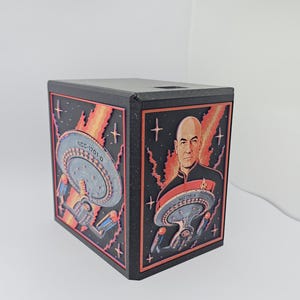 May include: A black rectangular box featuring raised, multi-colored relief panels on two sides. One side displays the Starship Enterprise NCC-1701-D in space. The other side shows a portrait of Captain Jean-Luc Picard alongside the starship.