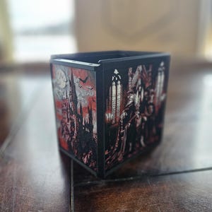 May include: Black, square container with a gothic-themed design. The artwork features a castle, bats, and figures in a dark red and gray color scheme. The container has a matte finish and is likely made of wood or a similar material.