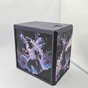 May include: A rectangular storage box with a dark, glittery frame. The front and side panels feature a textured, raised relief illustration of a monster in a black, purple, and blue cosmic nebula, with the creature breathing a stream of energy.