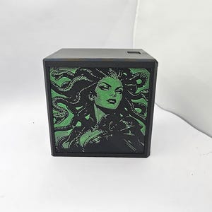 May include: A black square box featuring a graphic illustration of a woman with snakes for hair on the front panel. The artwork is rendered in shades of black and vibrant green, depicting a character with sharp features and a dark outfit.