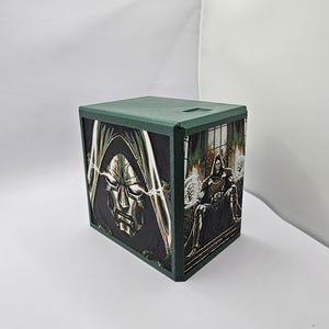 May include: A square green storage box featuring graphic illustrations of a masked character in a metallic hood and armor. The front panel displays a close-up of the character's face, while the side panel shows the character seated on a throne.