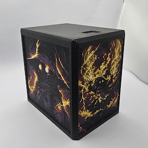 May include: A black rectangular box featuring two side panels with relief illustrations of a hooded figure with glowing yellow eyes. The artwork depicts the figure surrounded by swirling yellow and orange lightning effects against a dark background.