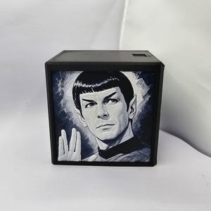 May include: A square black box featuring a monochromatic, grayscale illustration of a character with pointed ears and a bowl-cut hairstyle. The character is depicted performing a hand gesture with fingers parted in a V-shape, set against a dark background.