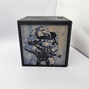 May include: A black square-shaped light box featuring a graphic print of a character in metallic gray power armor holding a large weapon. The armor has gold accents and the background is a textured gray. The frame is matte black plastic.