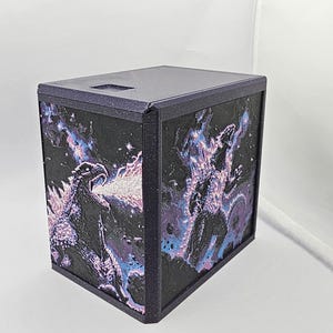 May include: A dark purple, rectangular storage box featuring a textured, three-dimensional relief of a monster breathing fire on the front and side panels. The relief design incorporates vibrant blue and pink highlights against a black background.