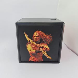 May include: A black square box featuring a printed image of a Klingon warrior with long hair holding a golden bat'leth weapon. The warrior wears a textured red and gold uniform. The box has a flat top surface with a small square cutout.