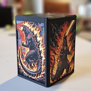 May include: Black, rectangular box featuring embossed images of Godzilla in fiery scenes. The artwork is in shades of red, orange, and black, with a textured appearance. The box is likely a collectible or decorative item.