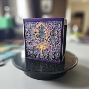 May include: A purple-framed, cube-shaped art piece with a textured, multi-colored front panel. The panel features a design with shades of black, purple, and yellow, possibly depicting a landscape or abstract scene. The cube sits on a black, circular base.