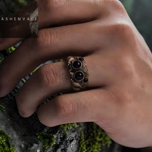 May include: A bronze-colored ring with four black gemstones. The ring has a textured surface and is worn on a finger. The ring's design is intricate and detailed, with a unique, artistic style. The ring is a piece of jewelry.