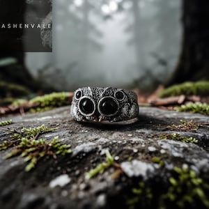 May include: A silver spider ring with large black gemstone eyes, set against a mossy rock with a blurred forest backdrop. The ring showcases detailed craftsmanship and texture. The Ashenvale logo is visible in the top left corner.