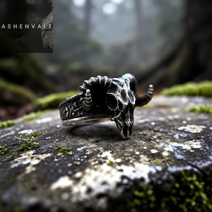 Może przedstawiać: Srebrny pierścień z motywem czaszki barana. Pierścień ma szczegółowe rzeźbienia na obręczy, a czaszka ma zakrzywione rogi. Pierścień jest umieszczony na naturalnym, mszystym tle. Słowo "ASHENVALE" jest widoczne w lewym górnym rogu.