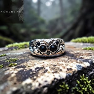 Puede incluir: Anillo plateado con un diseño texturizado detallado. El anillo presenta dos piedras negras redondas grandes, que parecen ojos, y piedras negras más pequeñas a cada lado. La palabra "ASHENVALE" es visible en la esquina superior izquierda.
