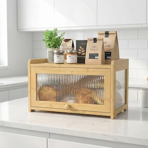 May include: A bamboo bread box with a clear, textured glass door, filled with loaves of bread. The top shelf holds jars, bags, and a small potted plant. The box is on a white countertop in a kitchen setting.