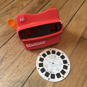 May include: A red View-Master 3D viewer with an orange lever and a "3D" label. A circular film reel labeled "Casper" is next to the viewer. The viewer has the words "View-Master" printed on the front.