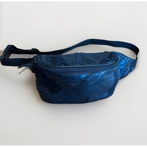 May include: A navy blue fanny pack with a patchwork design. The bag features a zippered main compartment and an adjustable strap. The leather fanny pack is a stylish accessory for carrying essentials.