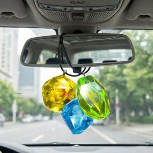 May include: Three faceted, jewel-shaped car charms in yellow, green, and blue hang from a black cord, suspended from a car's rearview mirror. The charms are transparent, catching the light.