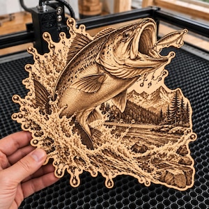May include: A laser-engraved wooden cutout featuring a detailed bass fish jumping out of water with a fishing lure in its mouth. The background depicts a mountain range with pine trees and rocky shores, surrounded by a decorative splash border design.