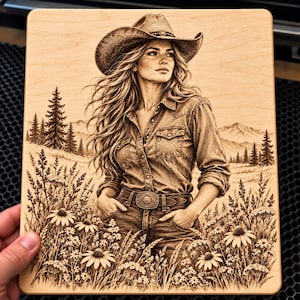 May include: A rectangular wooden plaque featuring a laser-engraved illustration of a person wearing a cowboy hat and a button-down shirt, standing in a field of wildflowers with pine trees and mountains visible in the background landscape.