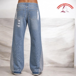 Peut inclure: Jean bleu clair avec une coupe large. Le jean pr&eacute;sente des d&eacute;tails d&eacute;chir&eacute;s et des &eacute;claboussures de peinture blanche. La vue arri&egrave;re montre les poches et la ceinture. Le logo "Versatile Fashions Art" est en haut &agrave; droite.