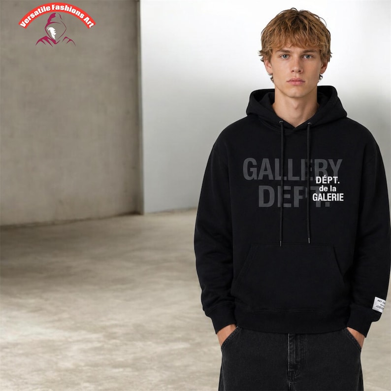 Puede incluir: Sudadera con capucha negra con el texto "GALLERY DEF" y "D&Eacute;PT. de la GALERIE" en gris. La sudadera tiene un bolsillo delantero y una capucha con cord&oacute;n. El modelo lleva vaqueros oscuros.