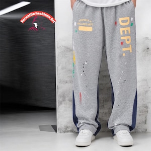 May include: Gray sweatpants with paint splatter details and the word "DEPT." in yellow. The pants have a blue stripe down the side and the text "PROPERTY OF GALLERY DEPT." in white. The model is wearing white sneakers.