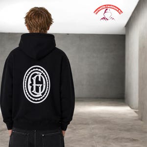 Puede incluir: Sudadera con capucha negra con un dise&ntilde;o ovalado blanco en la espalda. El dise&ntilde;o presenta una letra "G" estilizada dentro de un borde decorativo. La sudadera tiene capucha. Tambi&eacute;n se ve el texto "Versatile Fashions Art".