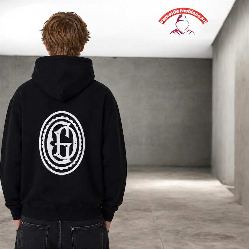 Puede incluir: Sudadera con capucha negra con un dise&ntilde;o ovalado blanco en la espalda. El dise&ntilde;o presenta una letra "G" estilizada dentro de un borde decorativo. La sudadera tiene capucha. Tambi&eacute;n se ve el texto "Versatile Fashions Art".