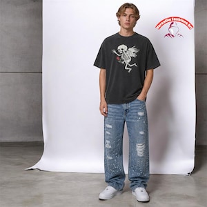 May include: A person wearing a dark grey t-shirt featuring a skeleton angel design and a red rose. The person is also wearing blue jeans with distressed details and white trainers. The image includes the text "Versatile Fashions Art".