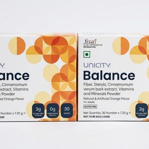 Op de afbeelding: Twee dozen Unicity Balance voedingssupplement. Elke doos is wit met abstracte oranje en gele ontwerpen. De dozen bevatten vezels, sterolen, kaneel, vitamines en mineralen. Ze zijn gelabeld met "Natuurlijke & Kunstmatige Sinaasappelsmaak" en "Glutenvrij".