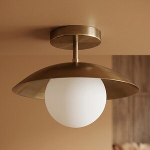 Könnte beinhalten: Eine Deckenleuchte in Messingoptik mit einer kugelförmigen weißen Glühbirne. Die Leuchte hat einen kreisförmigen Sockel und einen breiten, geschwungenen Schirm. Das Licht ist an einer neutralfarbenen Decke montiert.