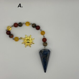 May include: A pendulum featuring a dark blue, faceted, cone-shaped stone suspended from a chain of alternating red, yellow, and speckled brown beads. A gold-colored sun charm is attached to the chain. The letter A is visible in the upper left.