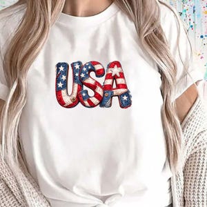 May include: White t-shirt with the word "USA" in a patriotic design. The letters are in red, white, and blue, with a patchwork style and star accents, reminiscent of the American flag. A casual, everyday wear item.