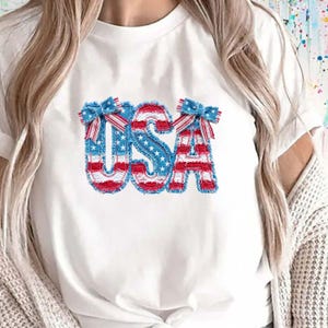 Op de afbeelding: Een wit T-shirt met korte mouwen en een grote afbeelding op de borst die het woord USA vormt. De letters hebben een gerafelde uitstraling met een rood, wit en blauw sterrenpatroon en twee kleine decoratieve strikjes.