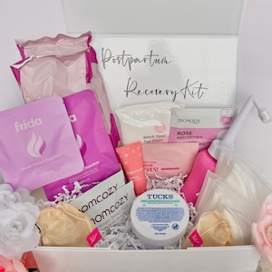 May include: A white box labelled "Postpartum Recovery Kit" filled with various items. Includes Frida heat relief patches, witch hazel pad liners, rose anti-oxygen facial mask, breast pads, and a peri bottle. The box contains a variety of pink and white products.