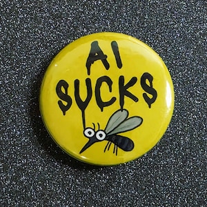 May include: A yellow button with the text "AI SUCKS" in black dripping font. A cartoon mosquito is drawn below the text. The button is round and has a glossy finish.