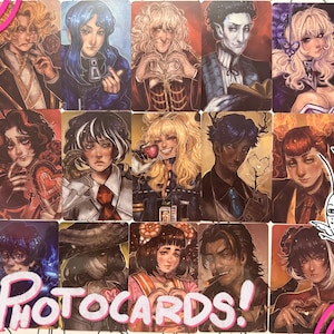 Può includere: Una collezione di fototessere con ritratti di personaggi in stile anime. Ogni carta presenta un personaggio diverso con acconciature, abiti ed espressioni uniche. Le carte sono rettangolari con uno sfondo chiaro e la parola "Photocards!" è scritta in rosa.