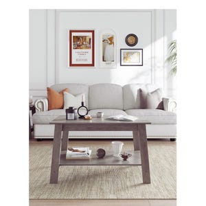 May include: A living room scene featuring a gray wooden coffee table with a lower shelf, holding books and decorative items. A light gray sofa with throw pillows sits behind the table. Artwork hangs on the white wall above the sofa.